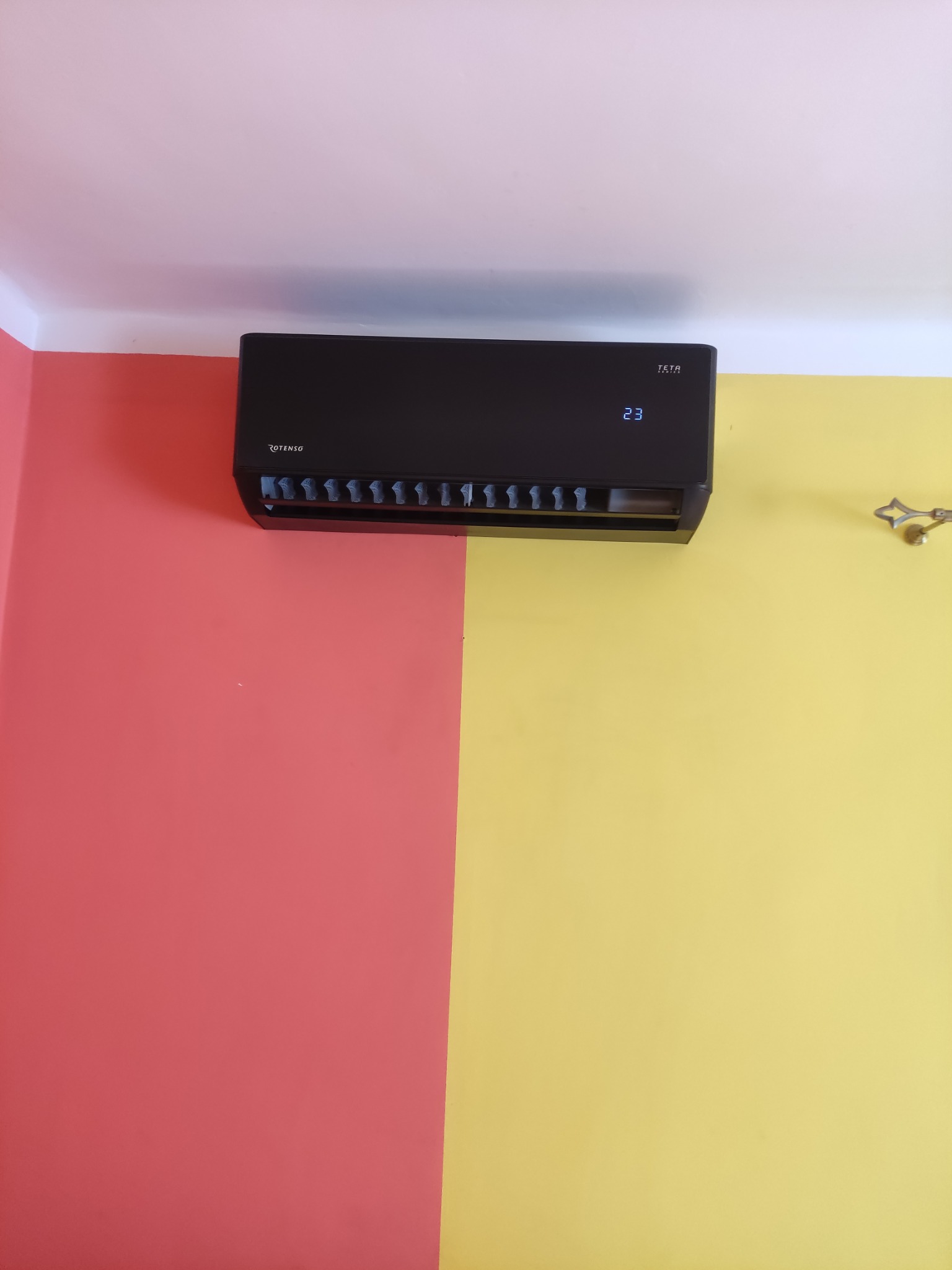 Czarny klimatyzator marki Rotenso na ścianie pomalowanej w kolorze żółtym i czerwonym, wyświetlający temperaturę 23 stopnie.