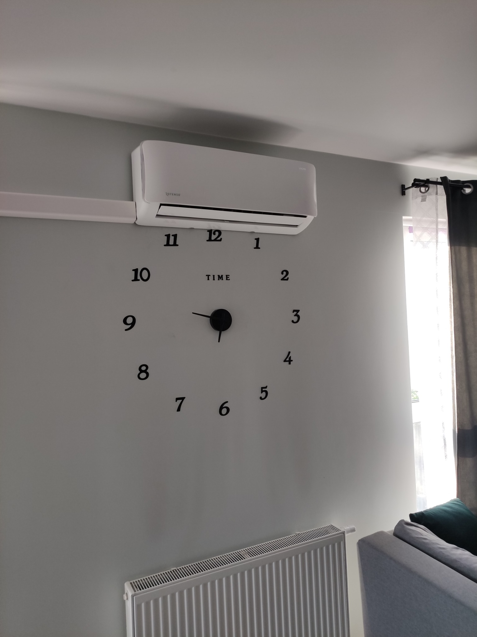 Biała jednostka klimatyzacji na ścianie nad dużym zegarem naklejonym na ścianę, widoczny grzejnik i fragment sofy