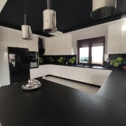 Nowoczesna kuchnia z białymi szafkami, czarnym sufitem i blatem, srebrnymi lampami wiszącymi i dekoracyjnym panelem z motywem limonki.