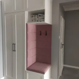 Biała szafa wnękowa z siedziskiem obitym r&oacute;żową tkaniną i wieszakami, uzupełniona p&oacute;łką z pudełkami, na tle szarej ściany i podłogi z płytek.