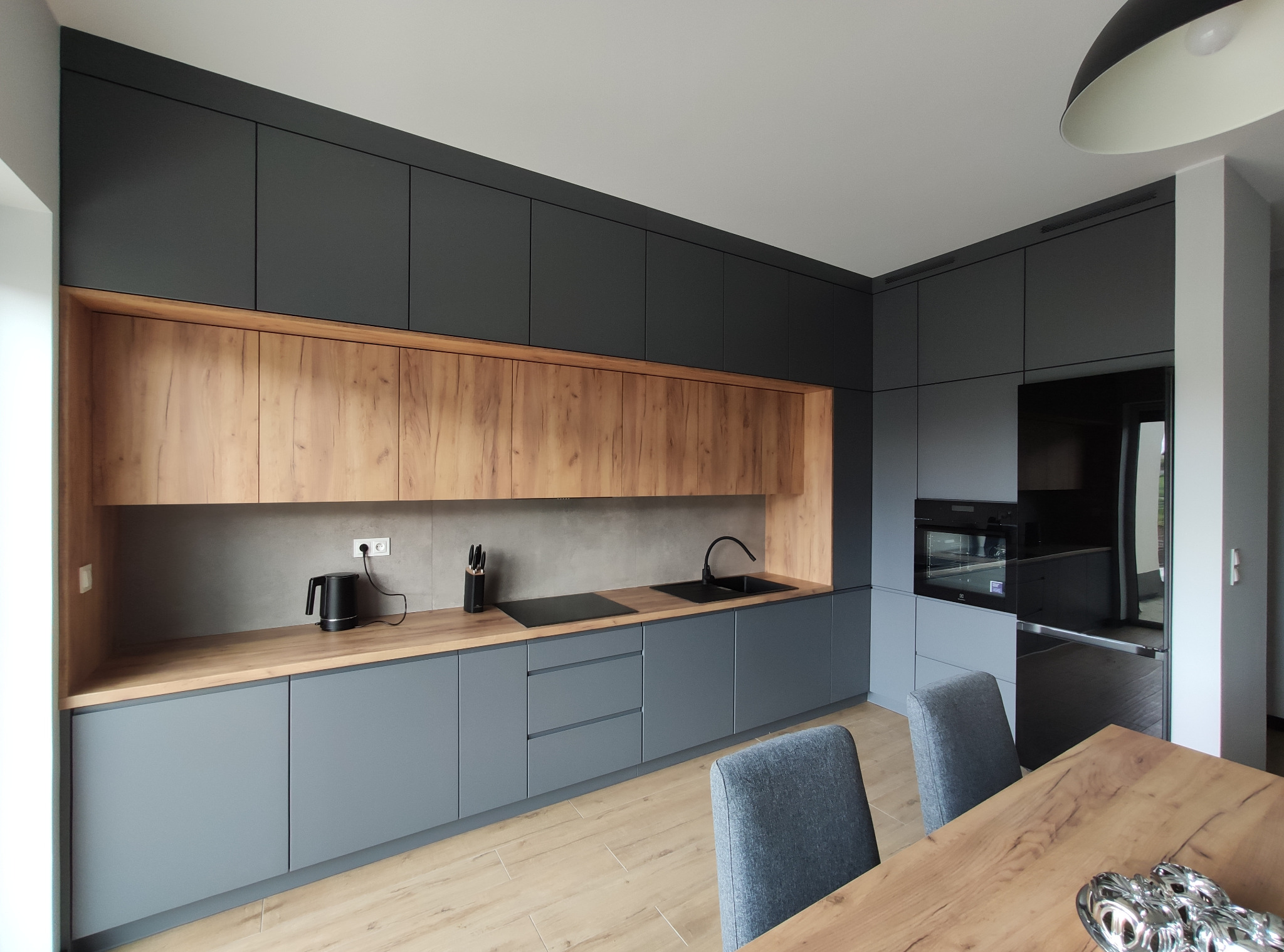 Nowoczesna kuchnia z szarymi szafkami dolnymi i drewnopodobnymi górnymi, czarny zlew i płyta grzewcza, drewniana podłoga i stół z szarymi krzesłami w tle.