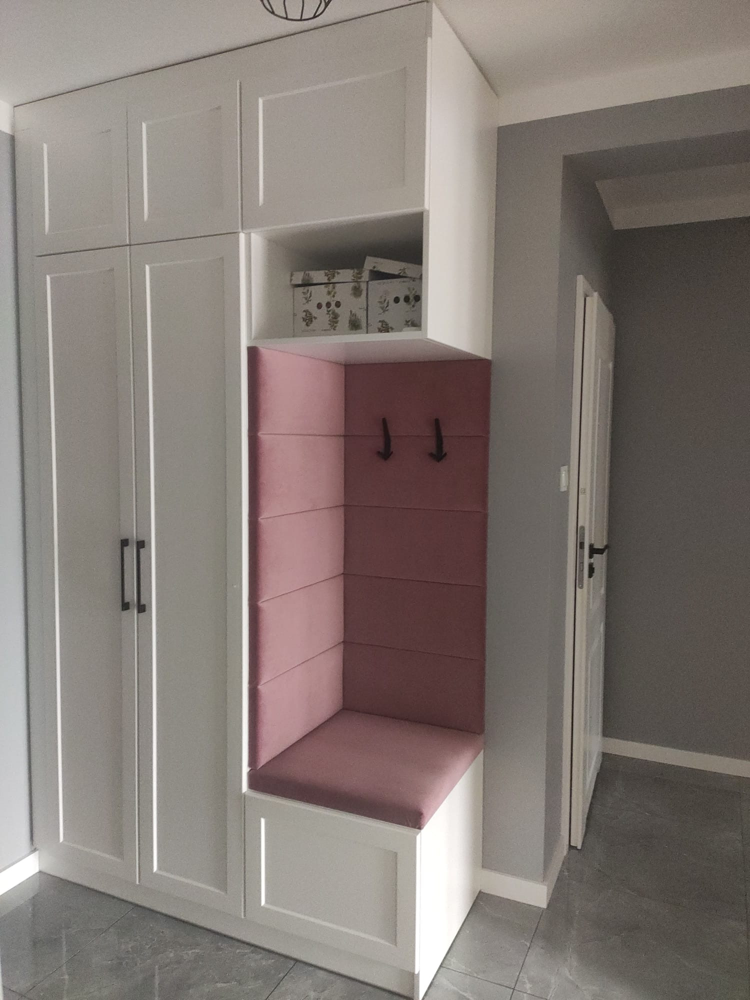 Biała szafa wnękowa z siedziskiem obitym różową tkaniną i wieszakami, uzupełniona półką z pudełkami, na tle szarej ściany i podłogi z płytek.