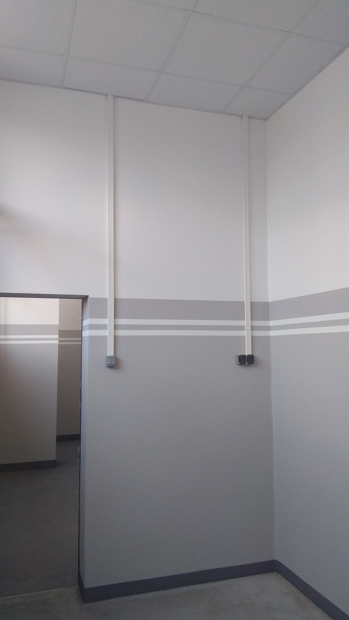 Wnętrze pomieszczenia z nową instalacją elektryczną: pionowe białe rury kablowe prowadzące od sufitu do szarych, podwójnych gniazdek elektrycznych na ścianie pomalowanej w poziome pasy szarości...