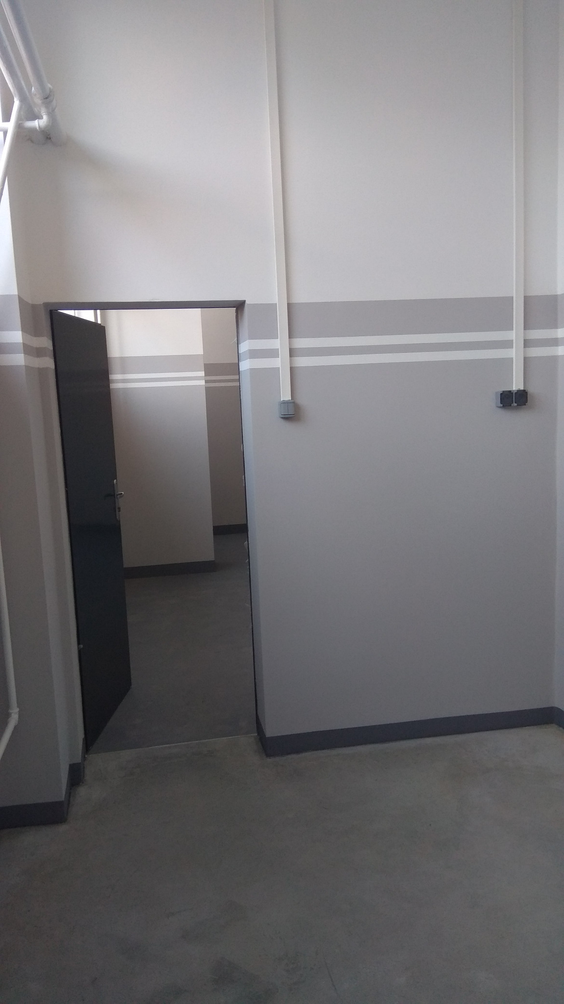 Szare pomieszczenie z otwartymi drzwiami, widoczne przewody elektryczne przy suficie i gniazdka na ścianie.