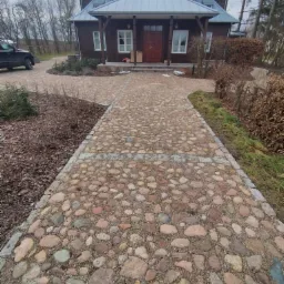 Kamienny chodnik prowadzący do brązowego domu z szarym dachem, otoczony zielenią i drzewami w pochmurny dzień.