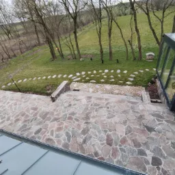 Kamienny taras z nieregularnych płyt przechodzący w schody i ścieżkę z kamieni prowadzącą przez trawnik, widok z góry na tle ogrodu i przeszklonej konstrukcji.