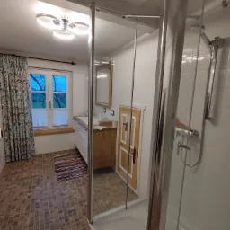 Jasna łazienka z kabiną prysznicową typu walk-in, drewnianą szafką pod umywalką i oknem z widokiem na zieleń, wykończona w neutralnych kolorach z akcentami w stylu rustykalnym.