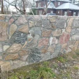 Niski mur z nieregularnych kamieni polnych w odcieniach szarości, brązu i czerwieni, spoinowany szarą zaprawą, na tle domu z ciemnego drewna i metalowym dachem.