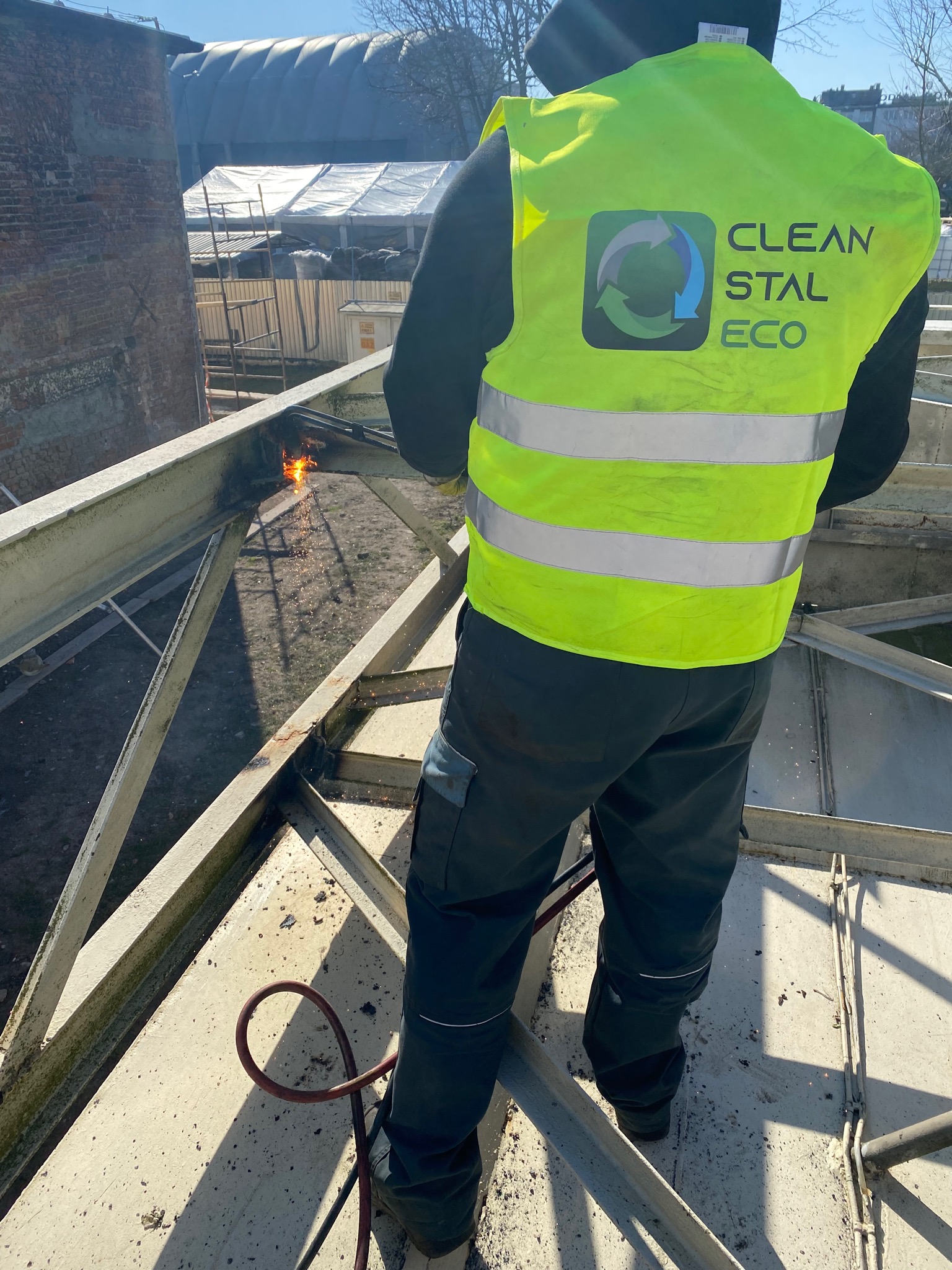 Pracownik w kamizelce odblaskowej z logo firmy Clean Stal Eco przecina metalową konstrukcję palnikiem, iskry lecą w dół, w tle budynek.