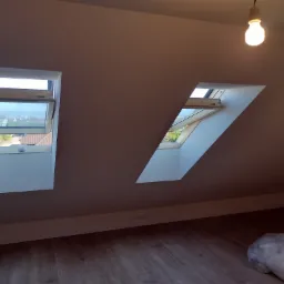 Poddasze z dwoma oknami dachowymi, jedno otwarte ukazujące widok na pobliskie domy, świeżo pomalowane ściany w odcieniach beżu, podłoga z paneli, wisząca żarówka i zakryty folią przedmiot w rogu...
