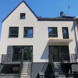 Nowoczesny dwukondygnacyjny dom z białymi ścianami elewacyjnymi, czarnymi ramami okiennymi i balkonami z metalowymi balustradami, widoczne schody wejściowe i fragment sąsiedniego budynku.