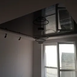 Błyszczący, czarny sufit napinany z wbudowanym oświetleniem punktowym i nowoczesną lampą wiszącą z okrągłymi elementami, odbijający widok z okna.