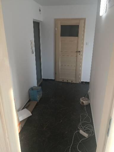 Wnętrze korytarza w trakcie prac wykończeniowych; widoczna zamontowana drewniana ościeżnica z ciemnym szybą, ciemna podłoga, kabel zasilający i pojemnik z farbą przy ścianie.