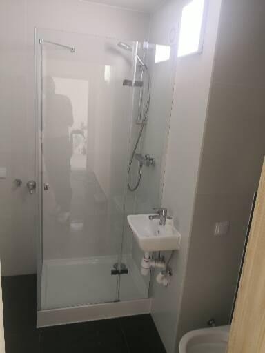 Nowoczesna kabina prysznicowa z pojedynczą szklaną ścianą, zamontowana umywalka z baterią, ciemna podłoga, widoczne elementy instalacji wodnej.