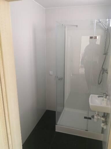 Nowoczesna kabina prysznicowa ze szklanymi drzwiami i małą umywalką narożną, czarne płytki podłogowe, białe ściany, minimalistyczny design.