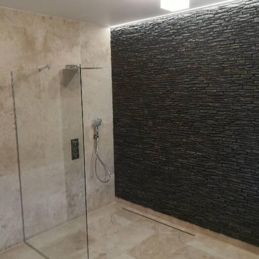 Nowoczesna łazienka z kabiną prysznicową ze szklaną ścianką, beżowymi płytkami imitującymi marmur i ścianą wyłożoną ciemnym, łupkowym kamieniem.