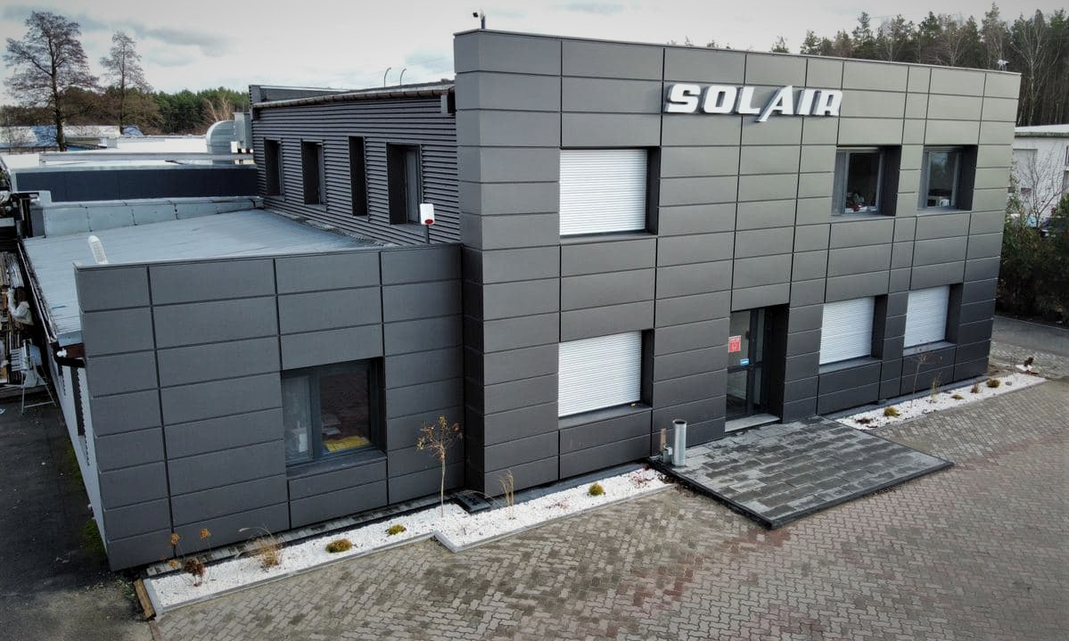 Nowoczesny budynek biurowy w szarym kolorze z logo firmy Solair na fasadzie, widok z zewnątrz na dwukondygnacyjny budynek z elementami wentylacyjnymi na dachu i roletami w oknach.