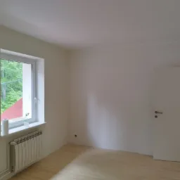 Pomalowane na biało wnętrze pokoju z drewnianą podłogą, grzejnikiem pod oknem i białą butelką z rozpylaczem na parapecie. Fragment drabiny malarskiej widoczny w lewym dolnym rogu.