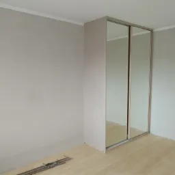 Świeżo zamontowana szafa wnękowa z przesuwnymi lustrzanymi drzwiami na panelach podłogowych w pokoju o pomalowanych na szaro ścianach. Na podłodze widoczne narzędzia montażowe.