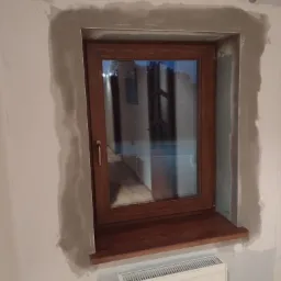Drewniane okno po montażu, z widocznymi śladami szpachlowania na ścianie i grzejnikiem pod parapetem.