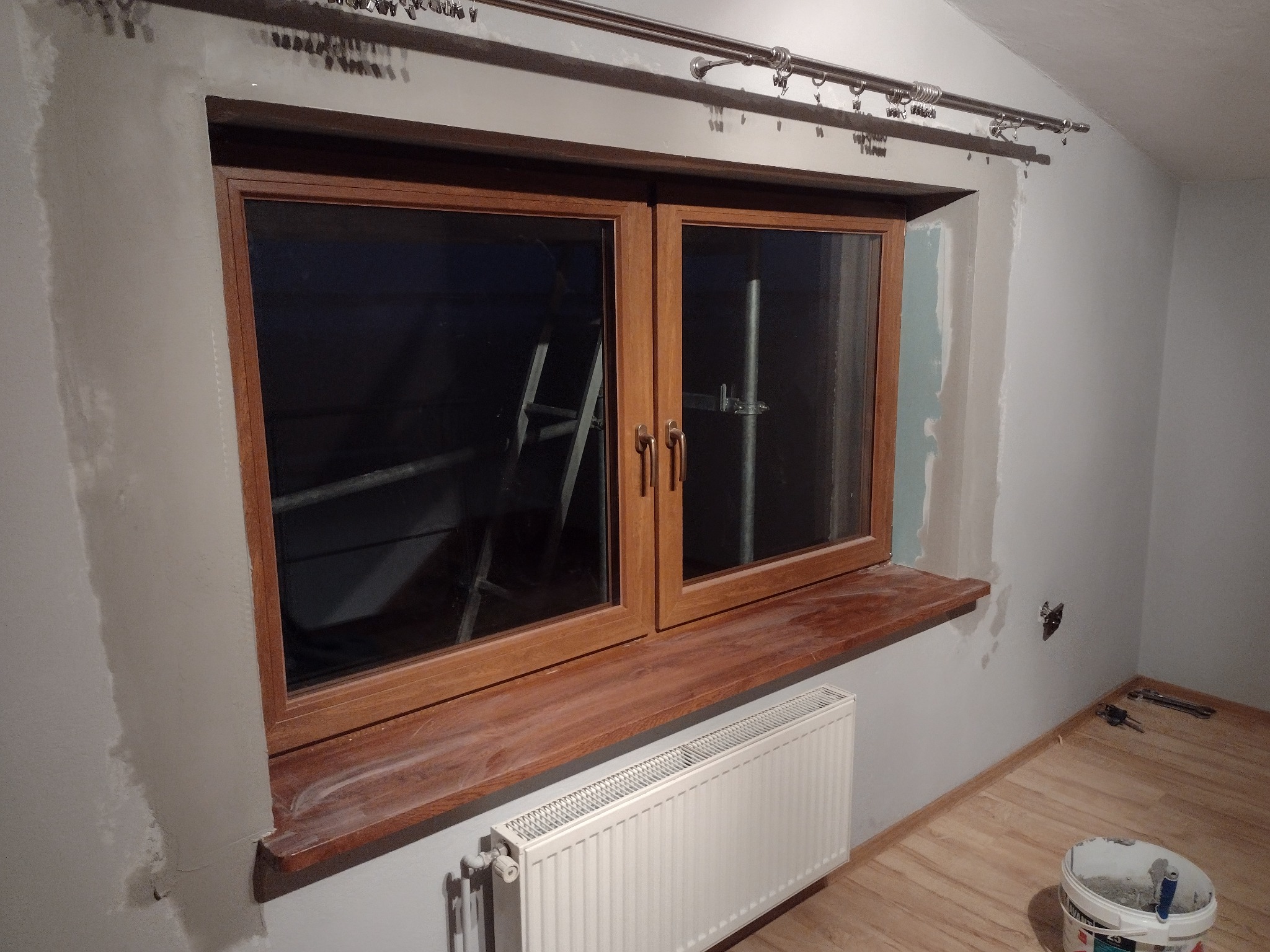 Przygotowanie do malowania pokoju: oszlifowane ściany, okno z drewnianym parapetem, grzejnik, wiadro z farbą, drabina w tle.