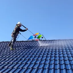 Pracownik w uprzęży, z kaskiem na głowie, czyści niebieski dach z dachówki ceramicznej za pomocą myjki ciśnieniowej; widoczne smugi wody na dachu.