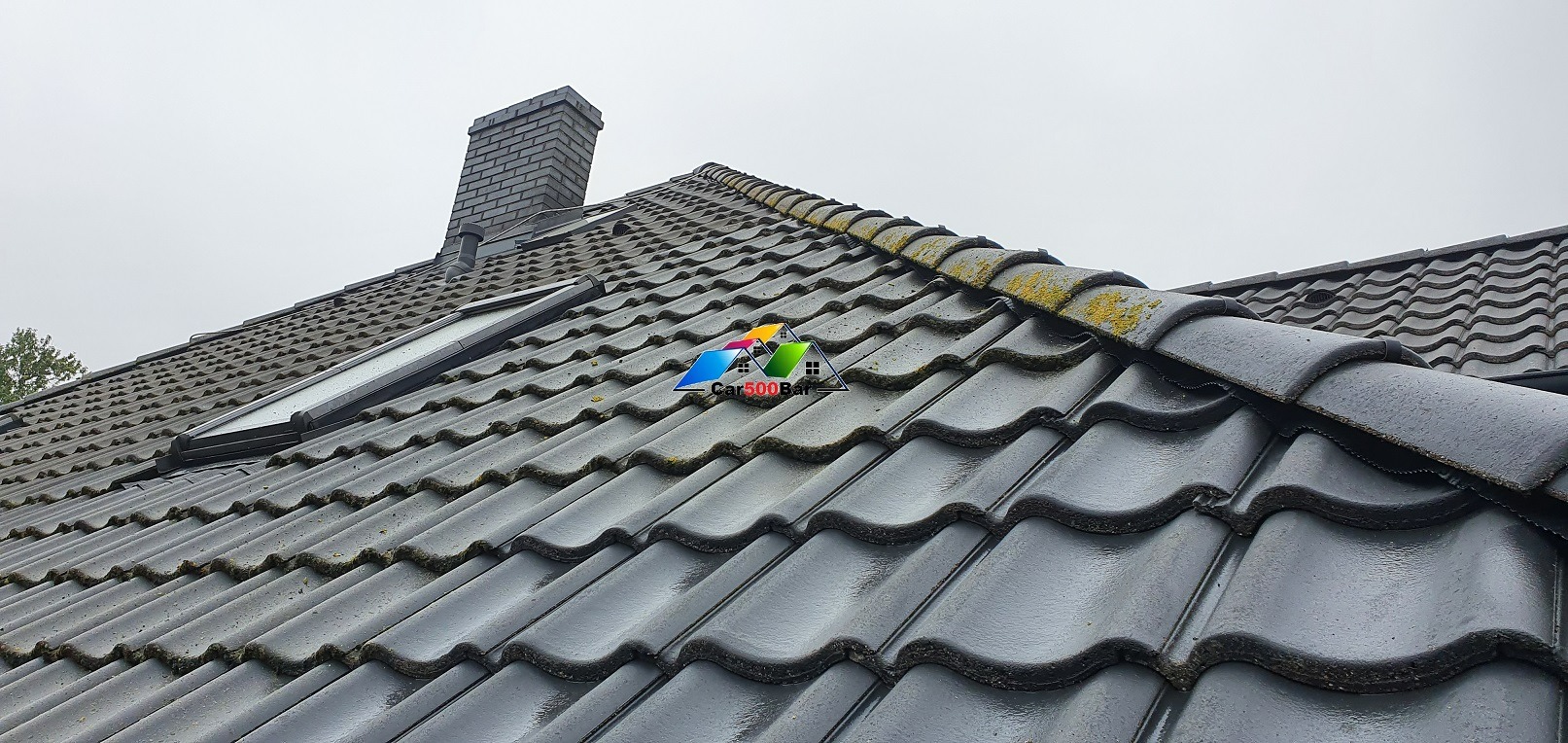 Ciemna dachówka ceramiczna z widocznymi śladami mchu i zabrudzeń, fragment komina z cegły oraz okno dachowe w pochmurny dzień.