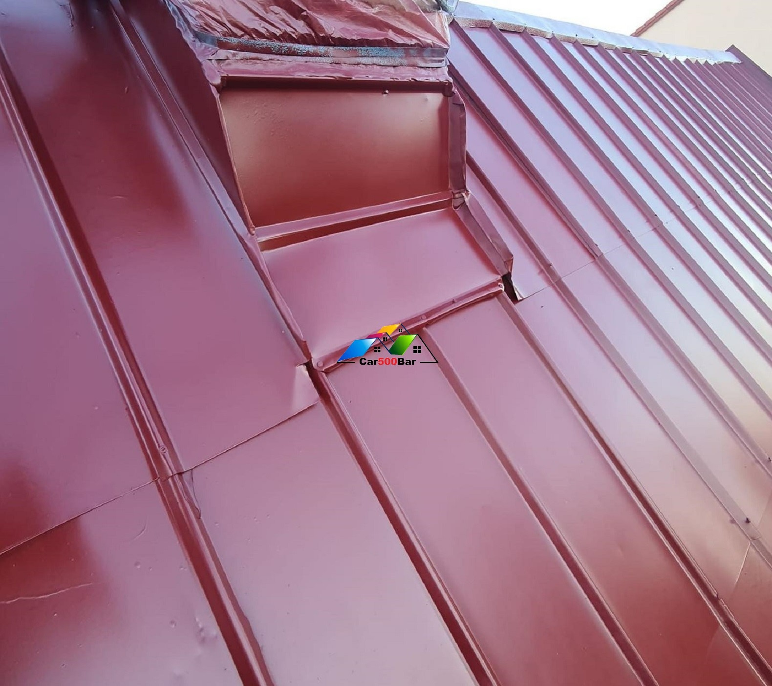 Błyszczący, bordowy dach z blachy trapezowej, z widocznym kominem obłożonym blachą. Część dachu pokryta folią ochronną. Widoczne połączenia blach.