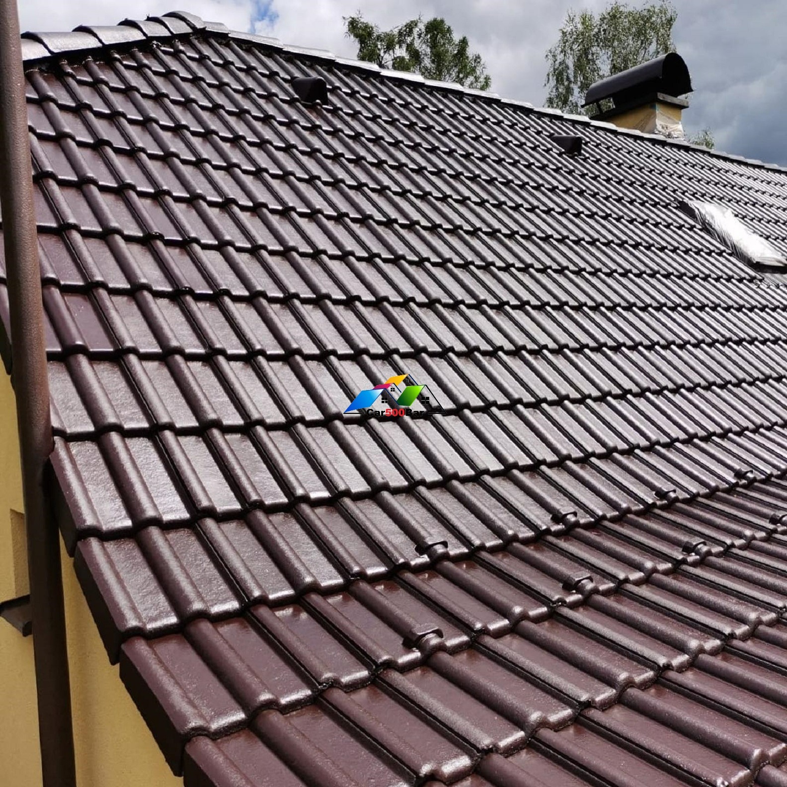 Błyszczący, brązowy dach z dachówki ceramicznej, widok z dołu pod kątem, z kominem i fragmentem rynny, po deszczu, z widocznymi kroplami wody.