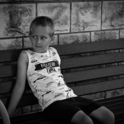 A. P. Foto - Chłopiec w koszulce z napisem 'Holiday Time' siedzi na drewnianej ławce na tle murowanej ściany, spoglądając w lewo.