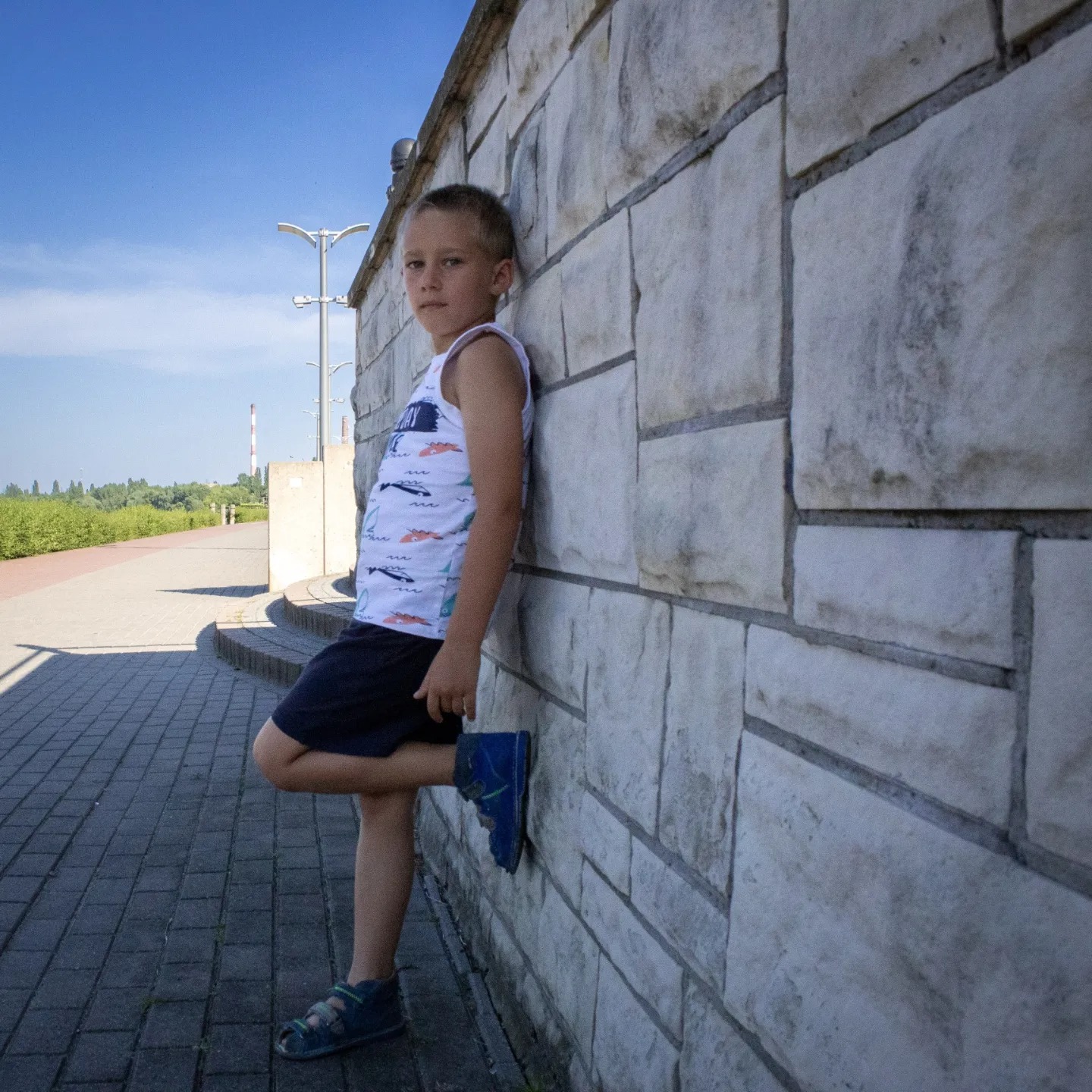 Chłopiec w koszulce z motywem ryb opiera się o kamienną ścianę na tle błękitnego nieba i zieleni.