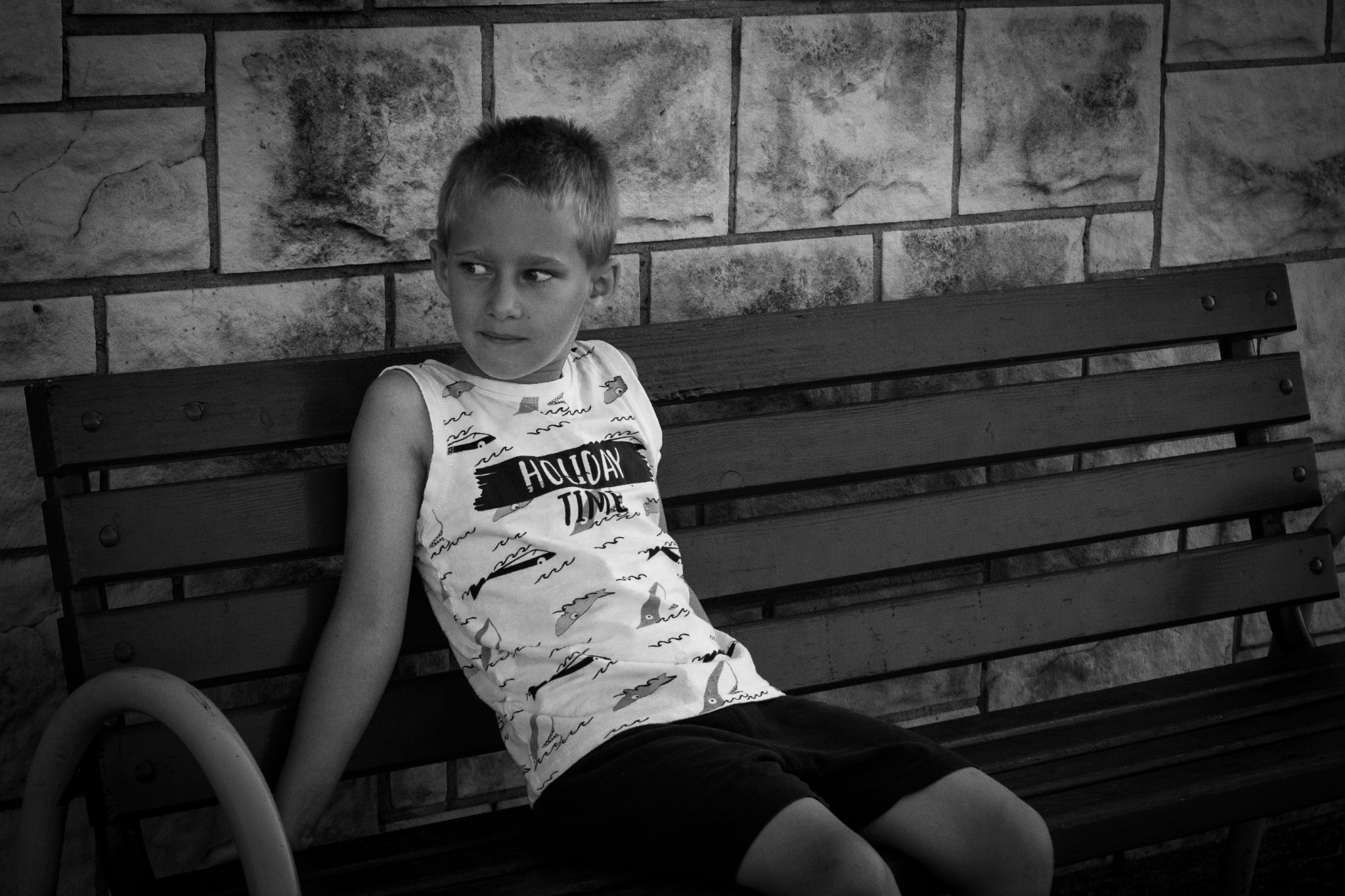 Chłopiec w koszulce z napisem 'Holiday Time' siedzi na drewnianej ławce na tle murowanej ściany, spoglądając w lewo.