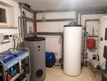 Kotłownia z trzema zbiornikami akumulacyjnymi ciepła: szarym z napisem TRINITY, białym cylindrycznym i niebieskim, instalacja rurowa pod sufitem, agregat prądotwórczy z przodu po lewej.