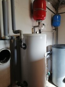 Biały bojler z podłączonymi rurami w szarej piance izolacyjnej, z czerwonym i niebieskim zbiornikiem wyrównawczym, widoczne elementy instalacji grzewczej w pomieszczeniu technicznym.