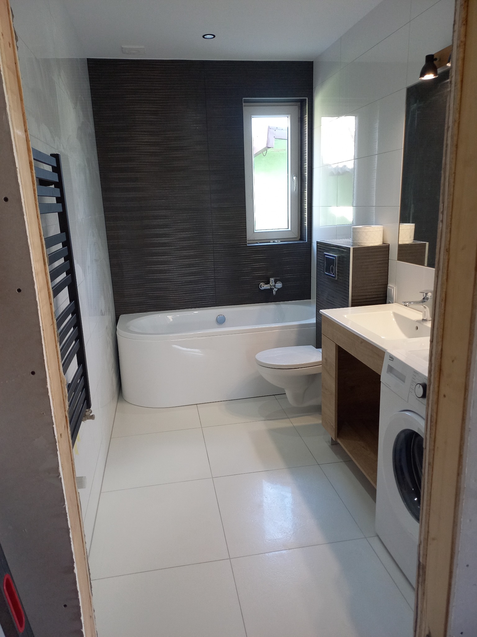 Nowoczesna łazienka po remoncie z białą wanną, umywalką z drewnianą szafką, pralką i grzejnikiem drabinkowym na tle ciemnych płytek imitujących drewno.