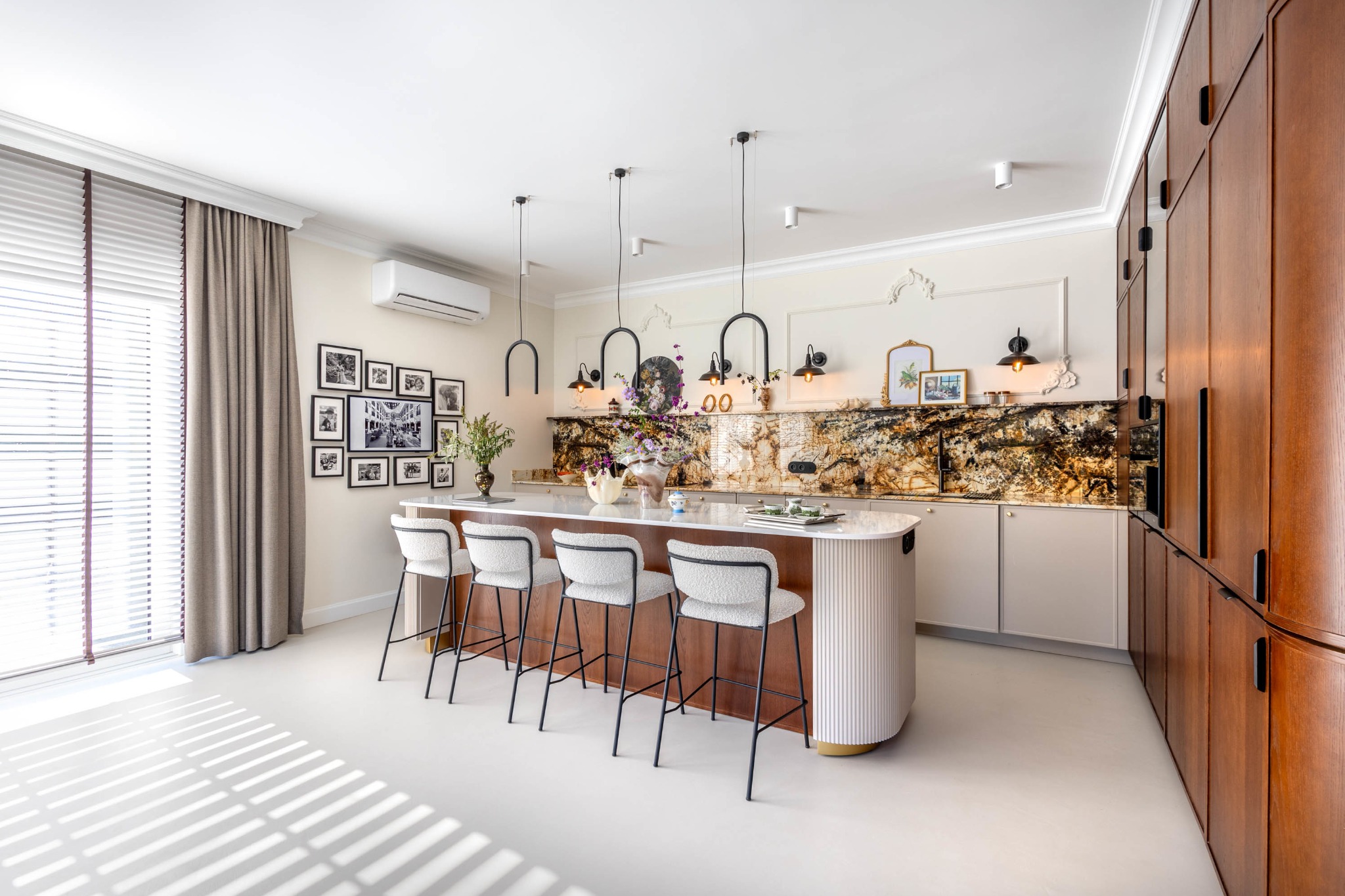 Nowoczesna kuchnia z wyspą i czterema krzesłami barowymi. Marmurowy backsplash, drewniane szafki, jasna podłoga i okno z żaluzjami. Dekoracyjne oświetlenie.