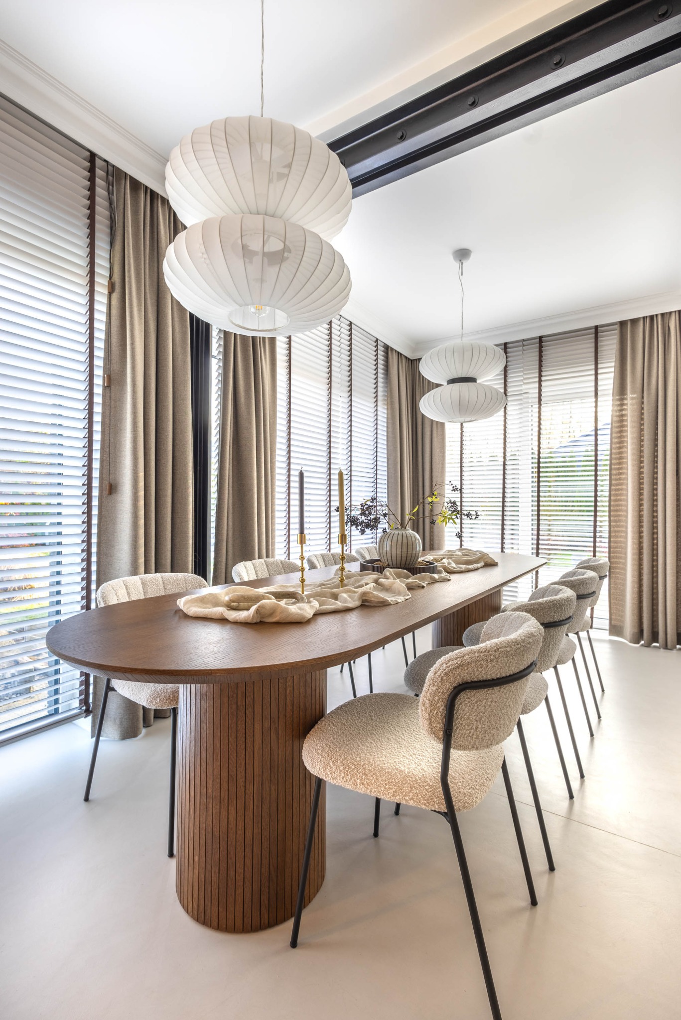 Stylowa jadalnia z owalnym stołem z drewna, krzesłami z boucle i lampami w stylu japońskim. Okno z żaluzjami i zasłonami w tle. Minimalistyczny wystrój.