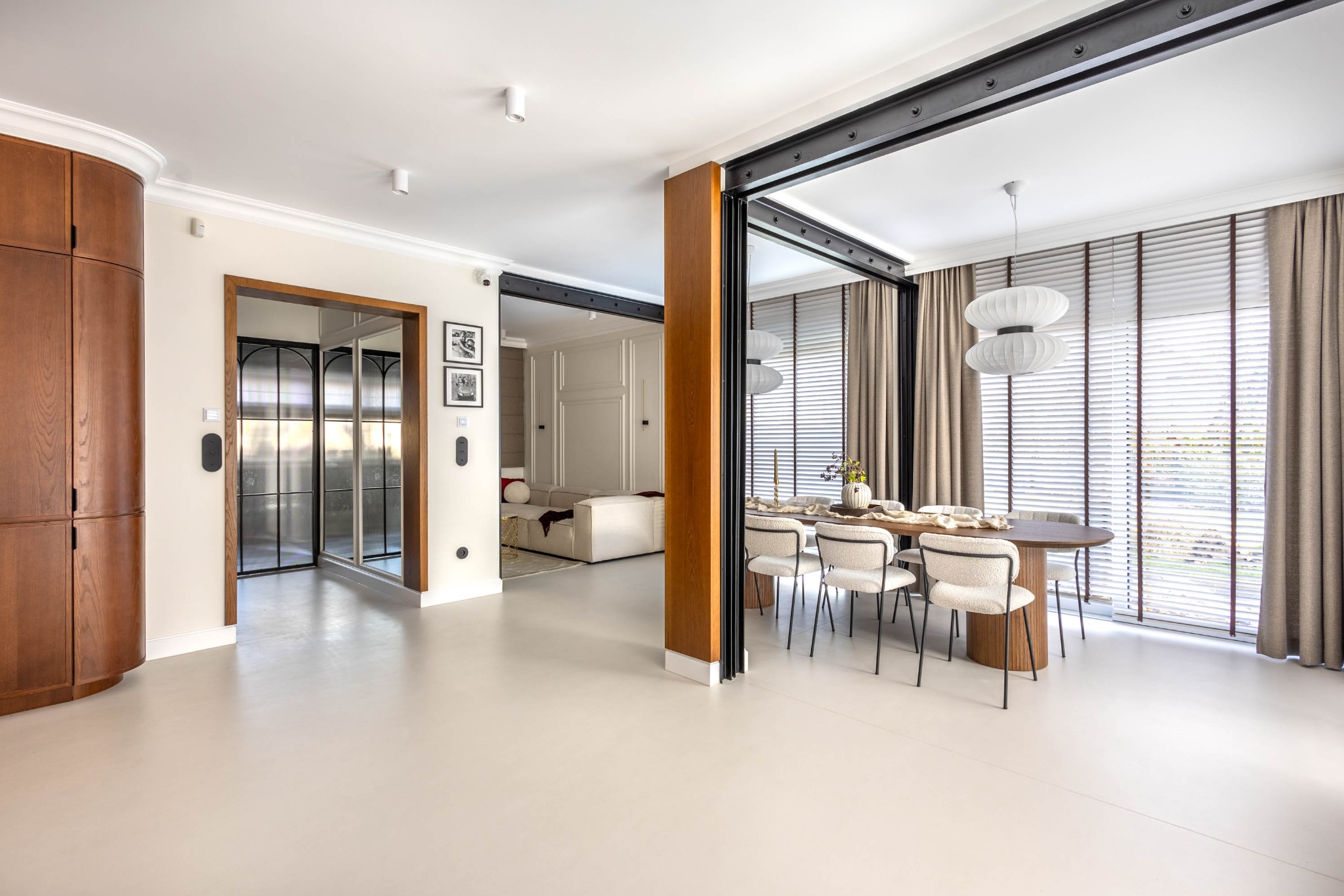 Jasne, przestronne wnętrze z drewnianą zabudową, stołem jadalnym i białymi krzesłami. Duże okna z żaluzjami i zasłonami wpuszczają naturalne światło. Minimalistyczny design.