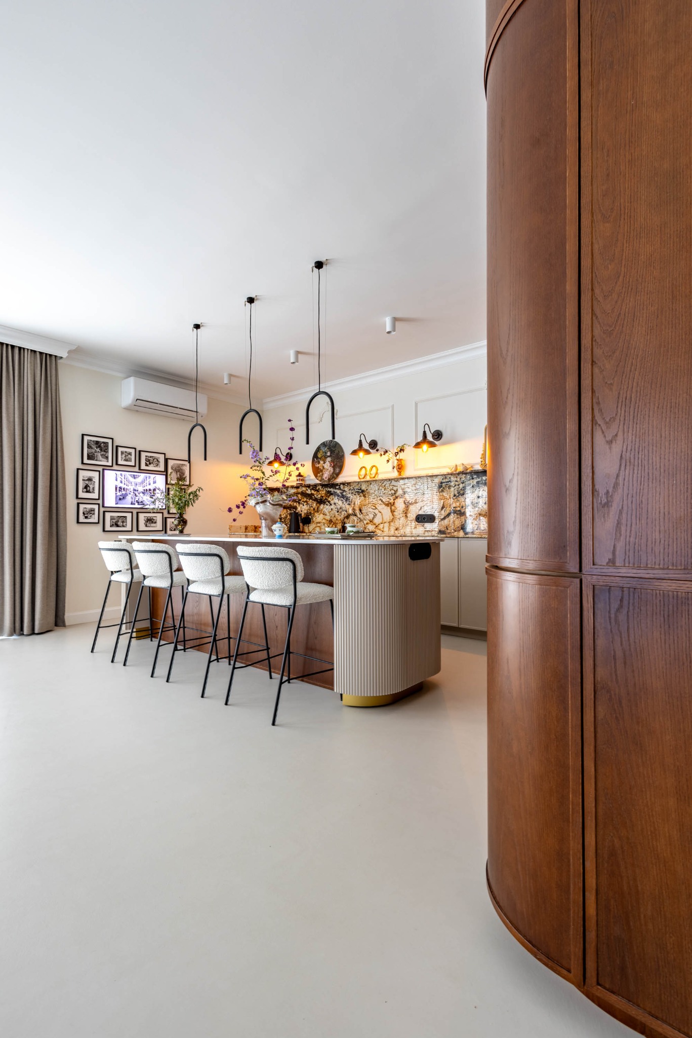 Stylowa kuchnia z wyspą i czterema krzesłami barowymi. Na ścianie galeria czarno-białych zdjęć. Ciepłe oświetlenie podkreśla marmurowy blat i dekoracyjny backsplash.