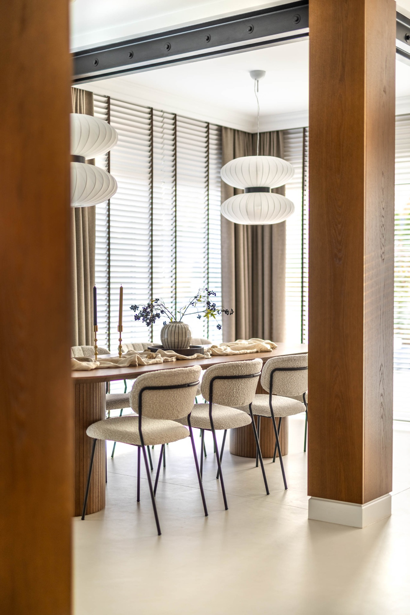 Elegancka jadalnia z drewnianym stołem, krzesłami z jasną tapicerką i dekoracyjnymi lampami. Widok przez drewniany filar, żaluzje w oknach i minimalistyczny wystrój.