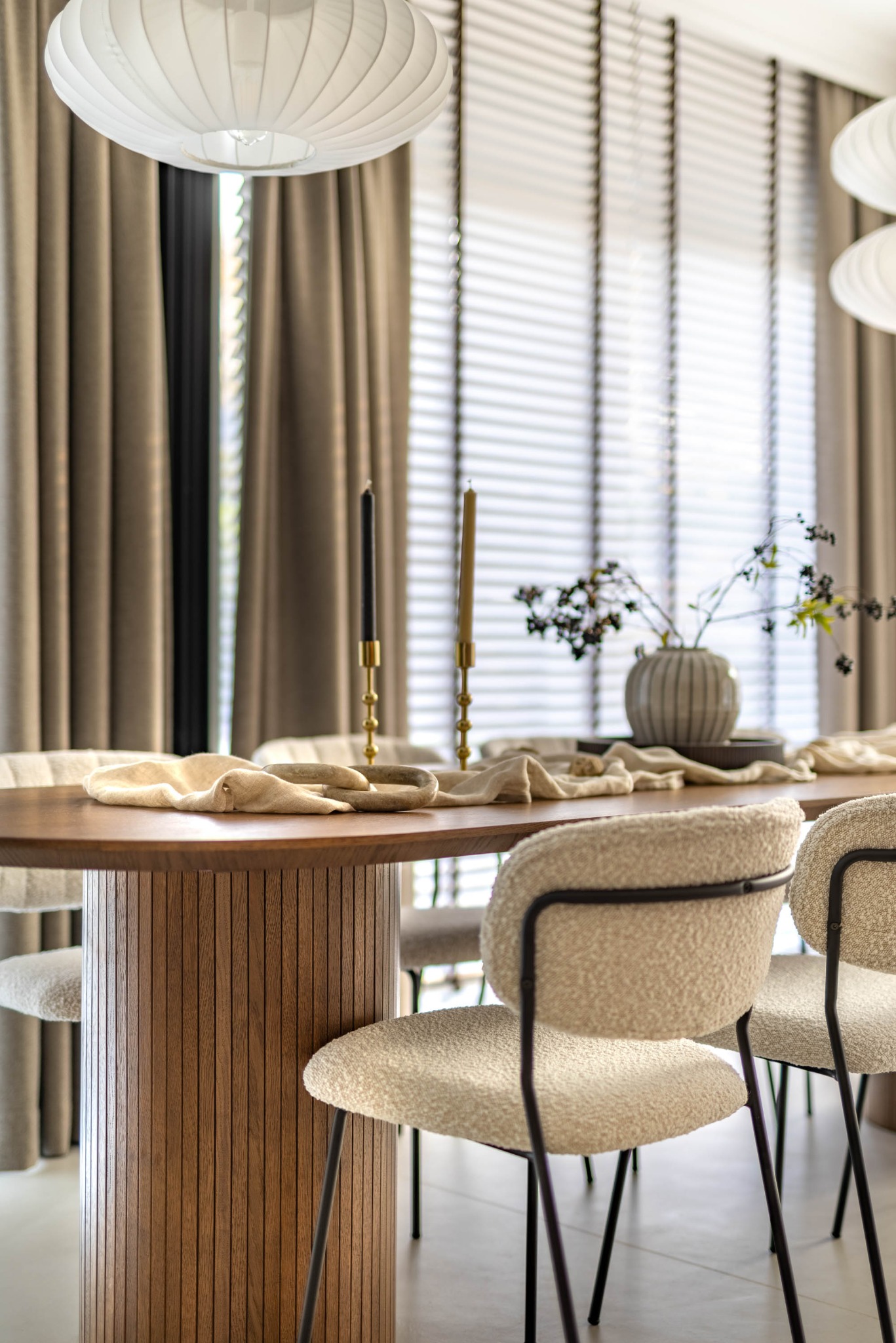 Elegancka jadalnia z drewnianym stołem, krzesłami boucle i dekoracjami: świeczniki, wazon z gałązkami. Stonowana kolorystyka, miękkie światło, żaluzje w tle.