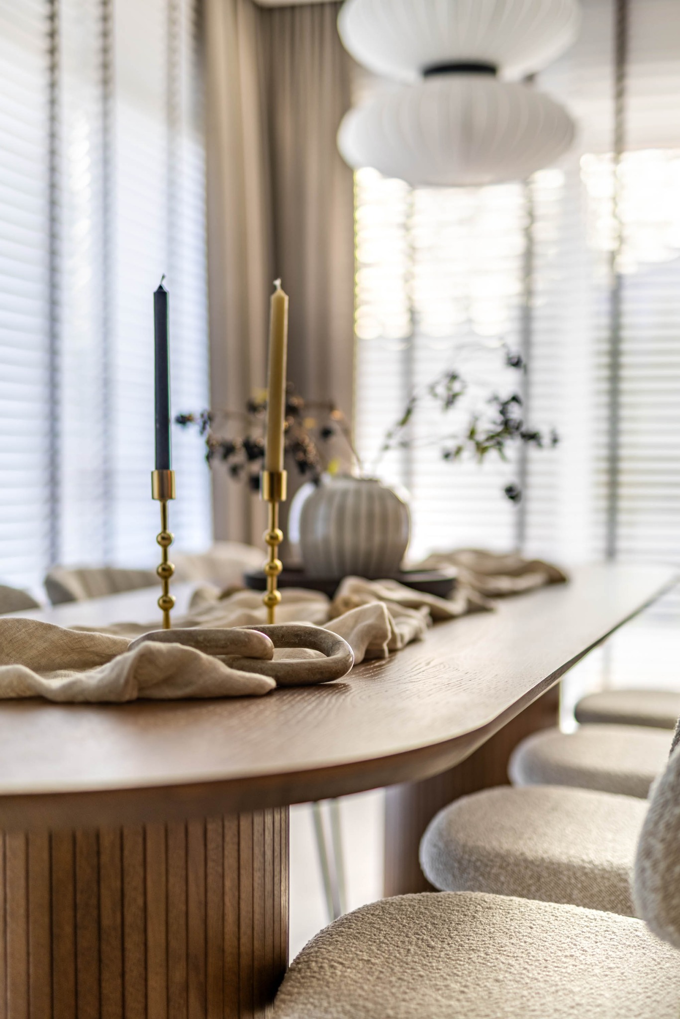 Stylowa aranżacja stołu w jasnym wnętrzu: drewniany stół z dekoracyjnym bieżnikiem, świeczniki z czarną i beżową świecą, wazon z gałązkami, miękkie krzesła i nowoczesna lampa.