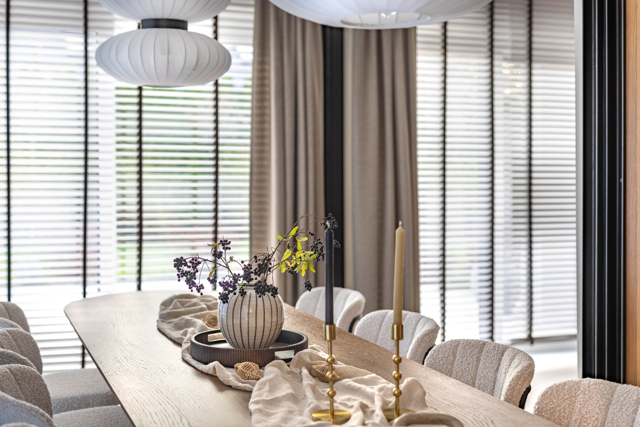 Stylowa aranżacja stołu w jadalni z drewnianym blatem, dekoracyjnym wazonem z gałązkami, świecami i tekstyliami. W tle okno z żaluzjami i designerskie lampy.