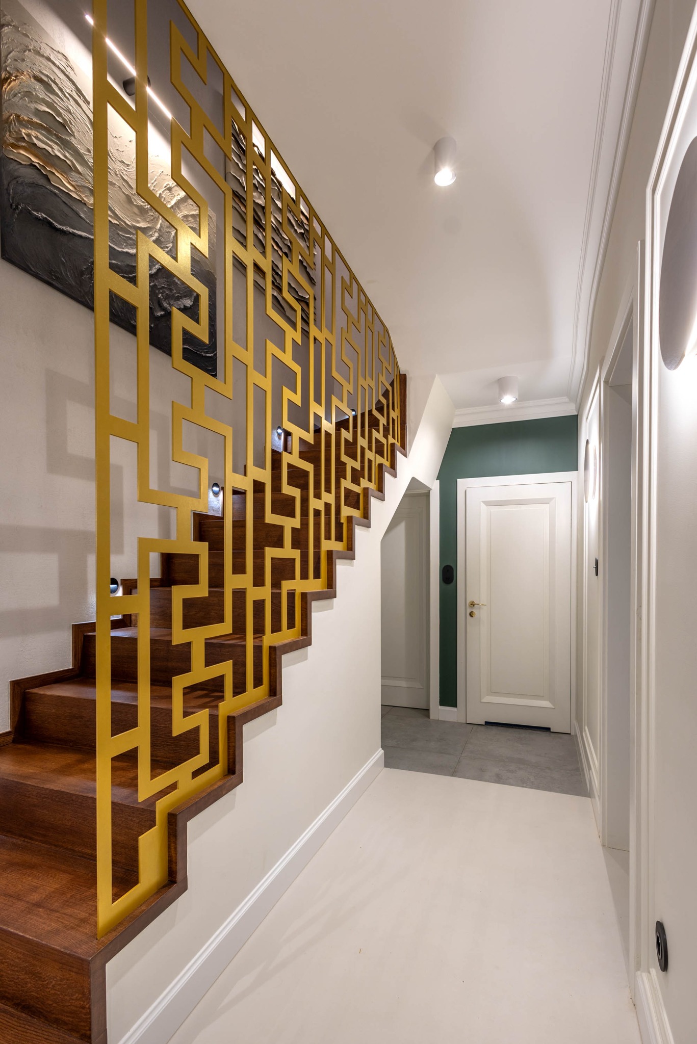 Nowoczesne schody z drewnianymi stopniami i złotą, geometryczną balustradą. W tle widoczny korytarz z białymi drzwiami i oświetleniem punktowym.