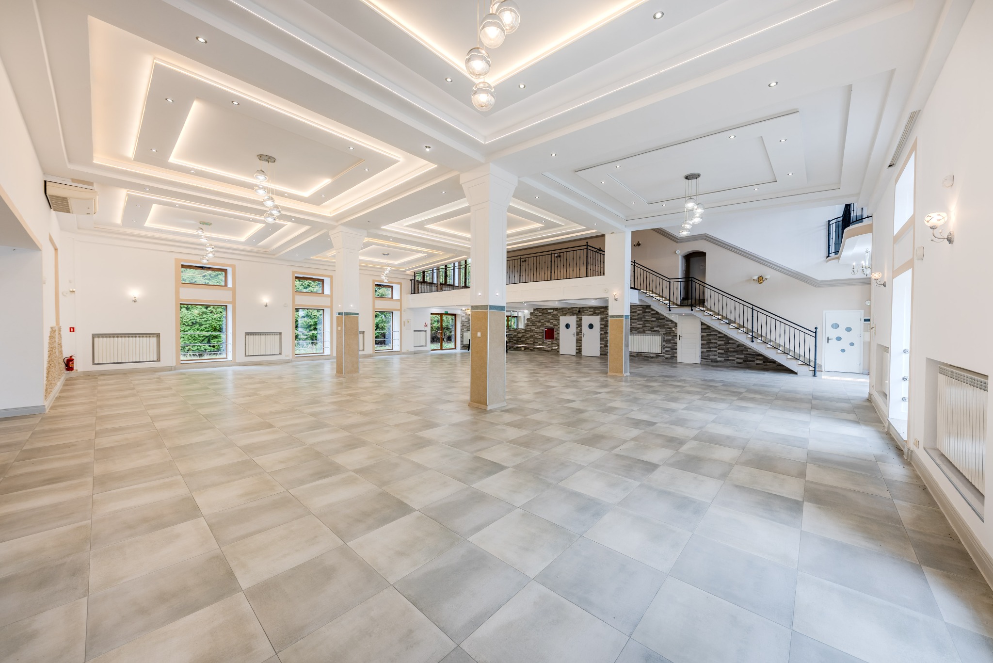Przestronne, jasne wnętrze sali balowej z kolumnami, schodami i dużymi oknami. Nowoczesne oświetlenie sufitowe i szara podłoga z płytek. Widok na zieleń za oknami.