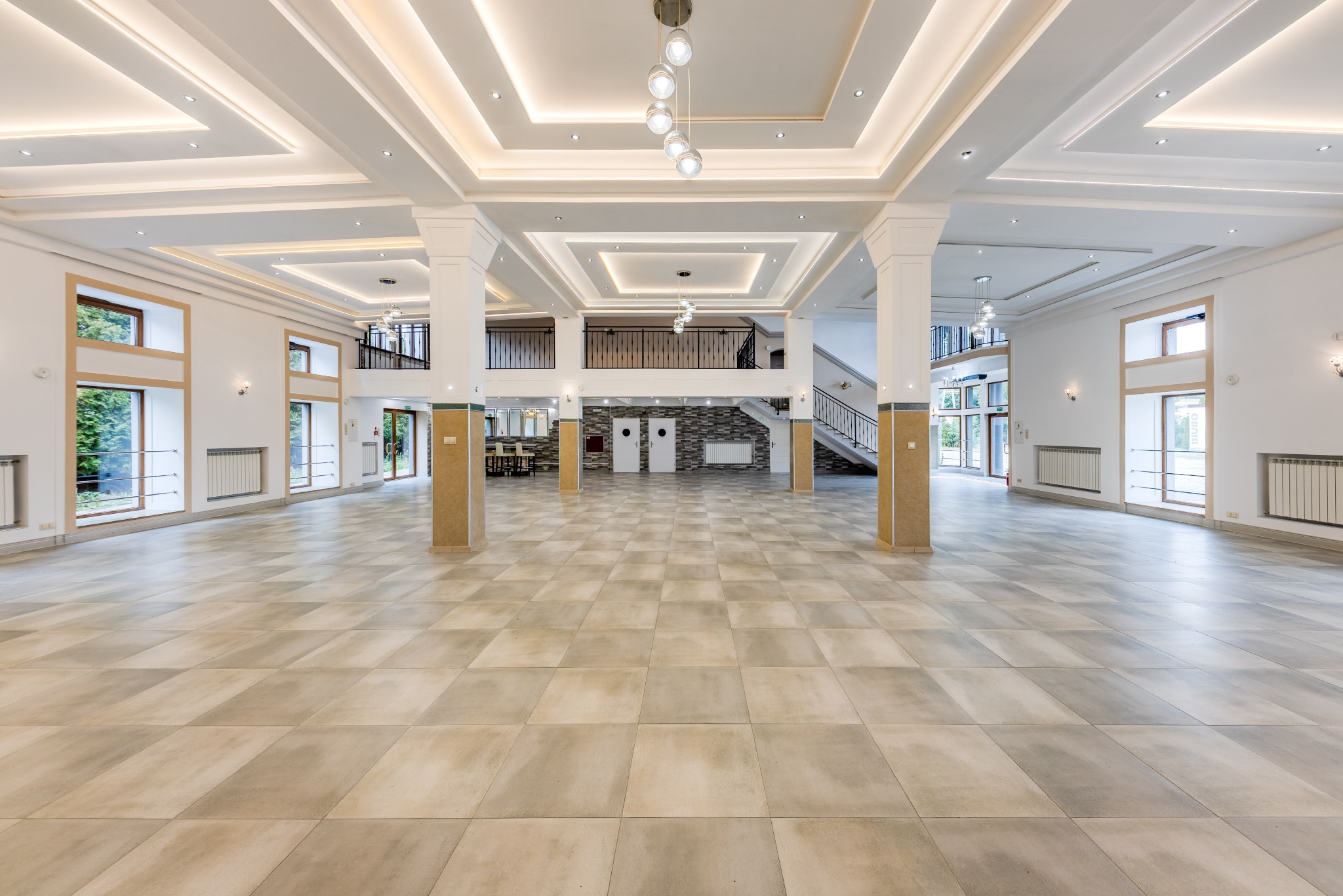 Przestronne, jasne wnętrze sali balowej z kolumnami, oświetleniem sufitowym i dużymi oknami. Parkiet w odcieniach beżu i szarości. Widoczne schody i antresola.