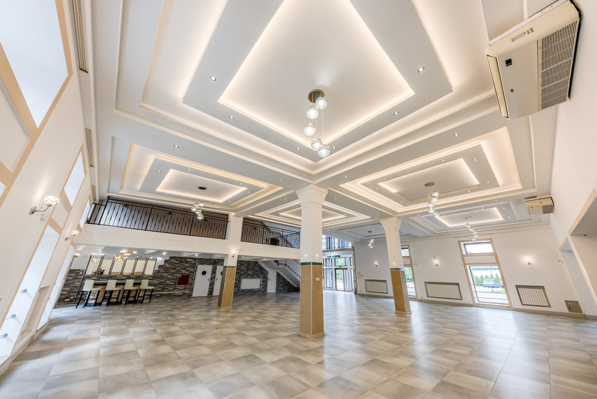Przestronne, jasne wnętrze sali balowej z antresolą, kolumnami i nowoczesnym oświetleniem sufitowym. Widoczne grzejniki pod oknami i bar z krzesłami w tle.
