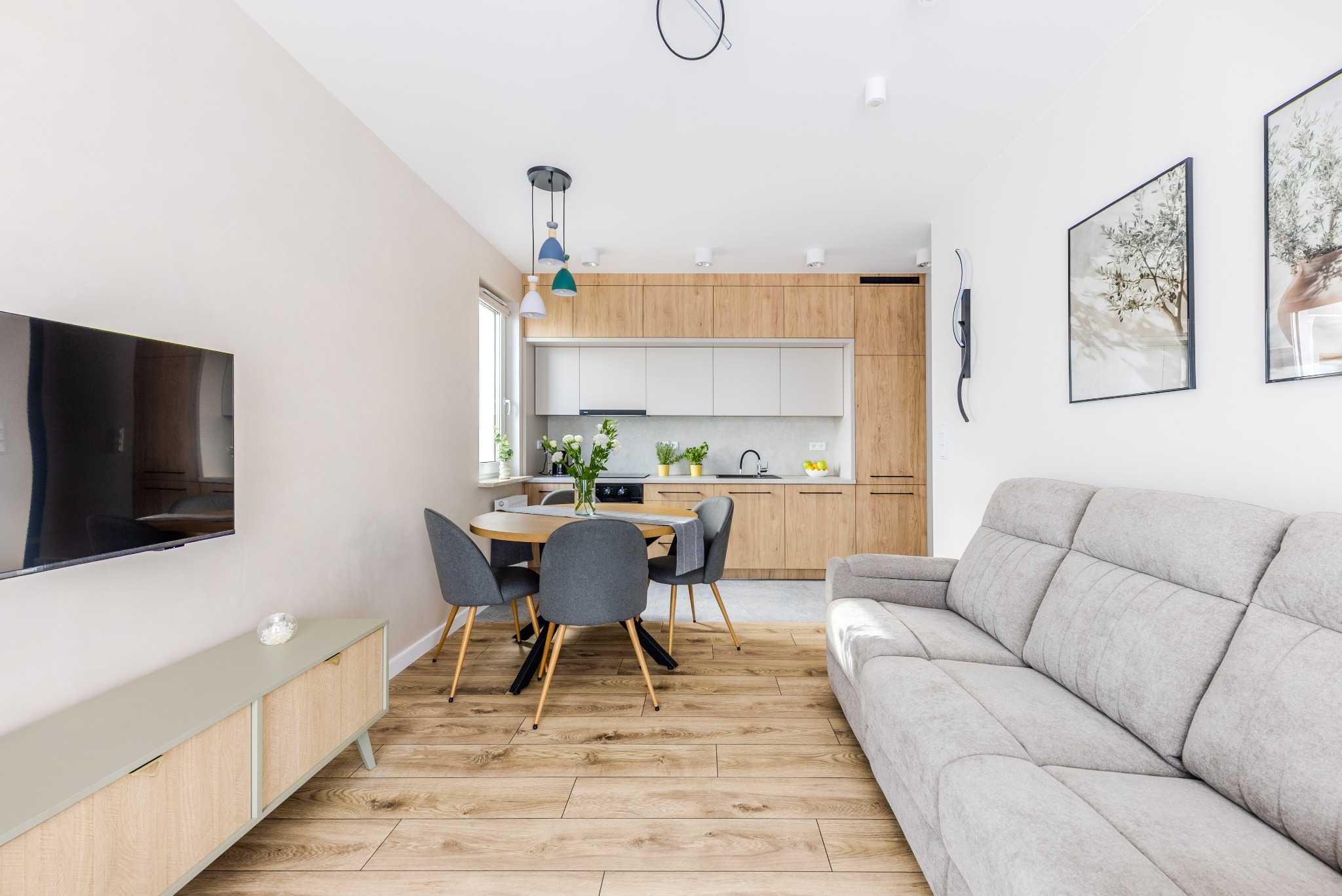 Jasne wnętrze mieszkania: drewniana podłoga, szara sofa, okrągły stół z krzesłami, zabudowa kuchenna z drewnianymi frontami. Minimalistyczny design, naturalne światło.