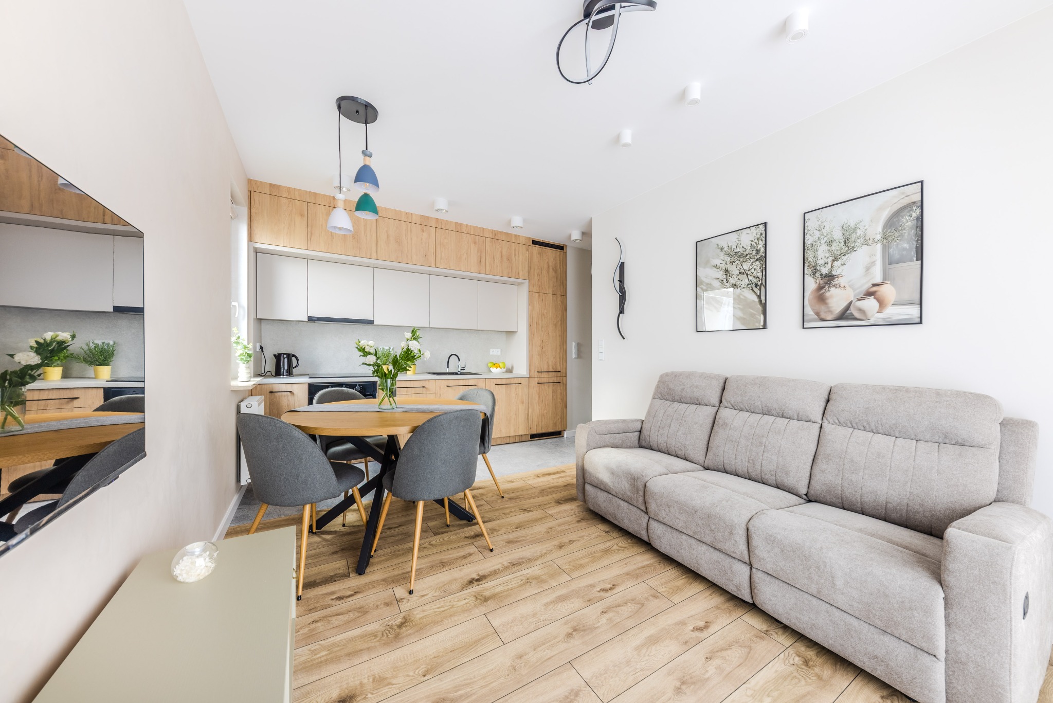 Jasne wnętrze: salon z aneksem kuchennym. Drewniana podłoga, szara sofa, okrągły stół z krzesłami. W tle zabudowa kuchenna z drewnianymi frontami i białymi szafkami.