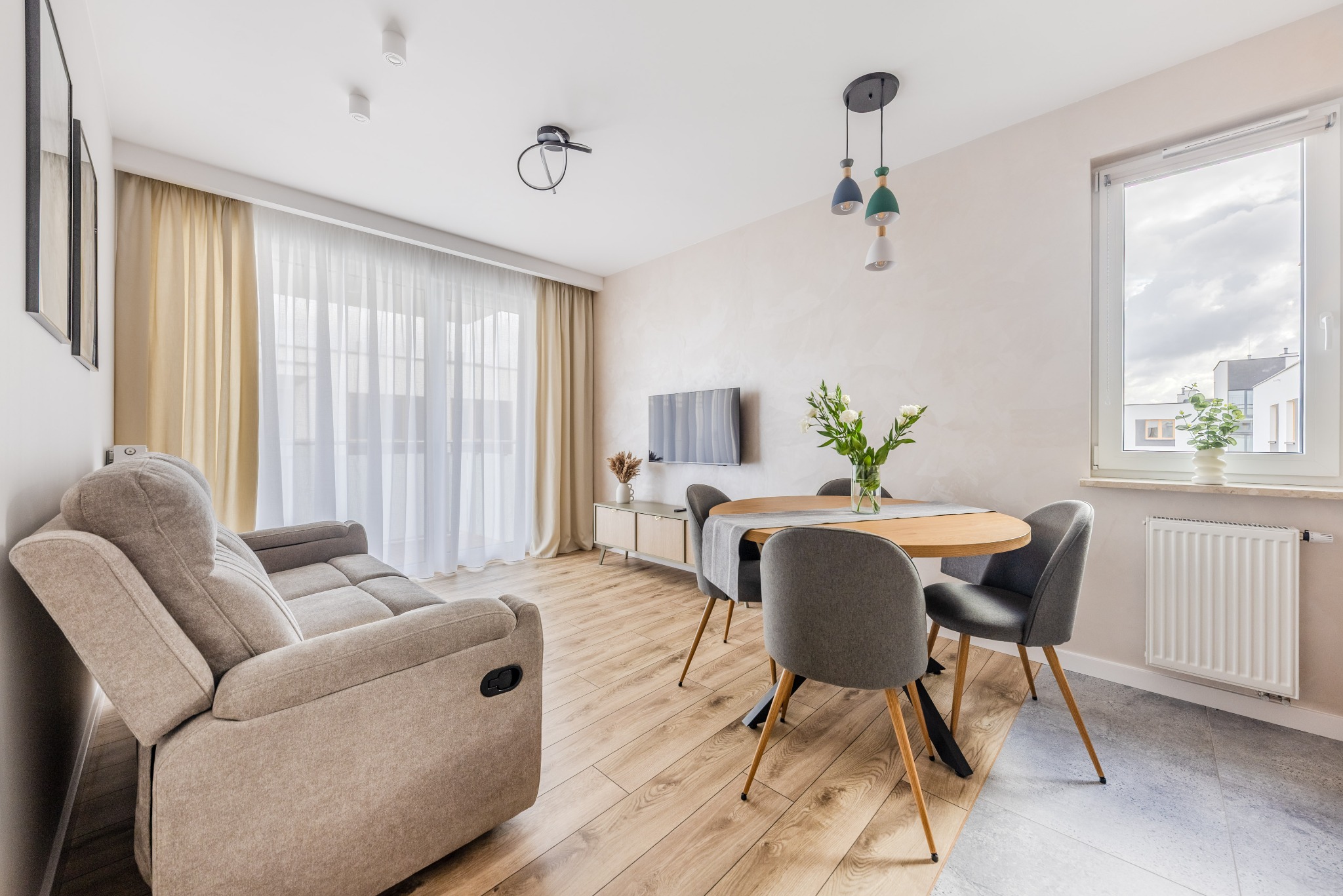 Jasne wnętrze salonu z drewnianą podłogą, szarą sofą z funkcją relaks, okrągłym stołem i kwiatami w wazonie. Nowoczesne lampy wiszące i telewizor na ścianie.